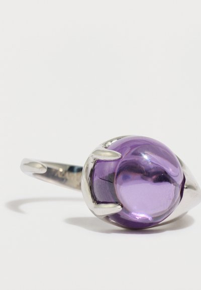 Ezüst gyűrű, amely nagy, sima, lila drágakövet tart karmokhoz hasonló foglalatban, polírozott felülettel és lekerekített formával.