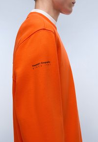 Felpa arancione con collo a giro, dettagli a coste e ricamo "Napapijri Geographic Since 1987" sulla manica.