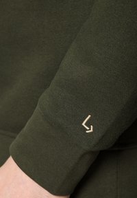 Ciemnozielony materiał bluzy o prążkowanej strukturze. Posiada małe, beżowe haftowane logo na mankiecie. Prosty i funkcjonalny design.