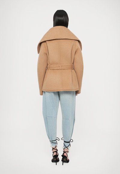 Egyenes sötét hajú nő öves tevebarna kabátban, világoskék, rojtos szegélyű farmernadrágban és fekete, fűzős, magas sarkú szandálban, háttal a kamerának.