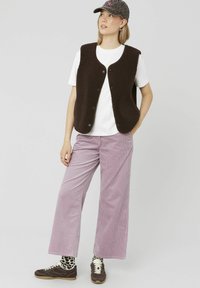 Un gilet en polaire marron porté sur une chemise blanche, associé à un pantalon en velours côtelé violet clair. Des baskets marron et des chaussettes à motifs complètent la tenue.
