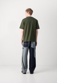 Olivgrünes Baumwoll-T-Shirt kombiniert mit Denim-Hosen im Patchwork-Stil, die verschiedene Blautöne und mehrere Taschen aufweisen, von hinten betrachtet.