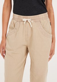 Beige Jogginghosen mit einem elastischen Bund, strukturiertem Stoff, seitlichen Taschen und einem dezenten Rippendesign entlang des Bundes und der Taschen.