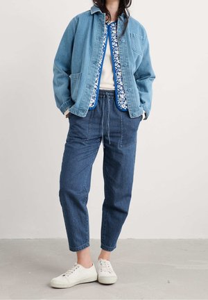 Personne portant une veste en jean bleu clair, un gilet fleuri bleu, un jean bleu foncé avec taille à cordon et des baskets blanches, debout à l'intérieur.