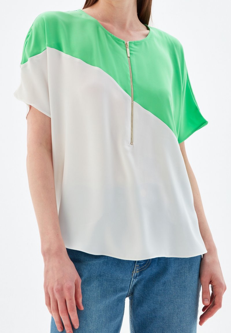 Haut à manches courtes en vert et blanc, présentant un design en bloc de couleur diagonale. Tissu lisse avec un détail de fermeture éclair à l'avant.