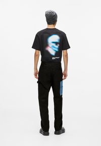 Tricou negru din bumbac cu un design grafic colorat pe spate, combinat cu pantaloni cargo negri cu o petec din denim albastru. Beanie negru și pantofi.