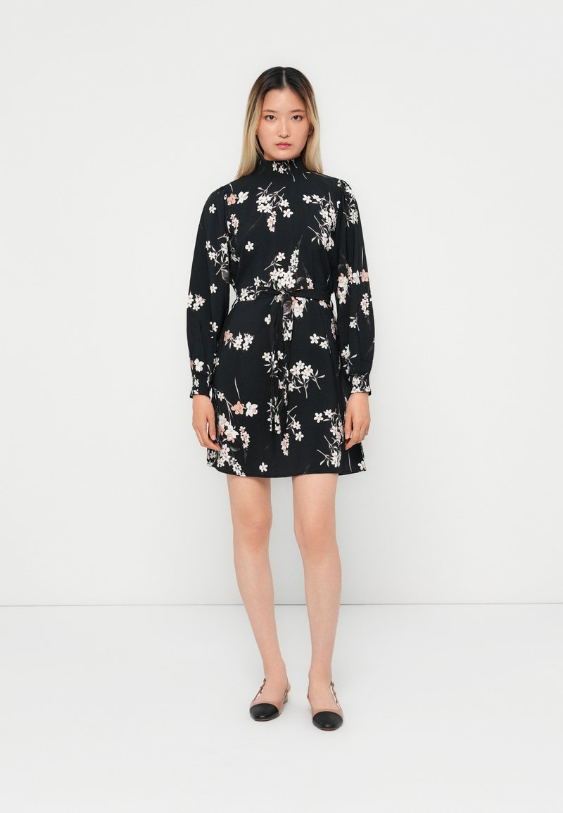Vestido preto de mangas longas, estampa floral em rosa e branco, cintura cingida, comprimento até aos joelhos, usado com sapatos pretos rasos.
