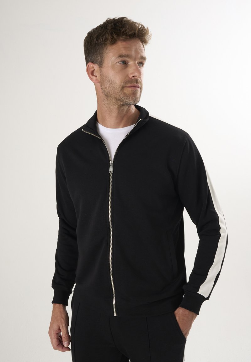 Svart zip-up jacka med hög krage, med vita sidostreck och en silverfärgad dragkedja. Tillverkad av mjukt tyg, långärmad och med en avslappnad passform.