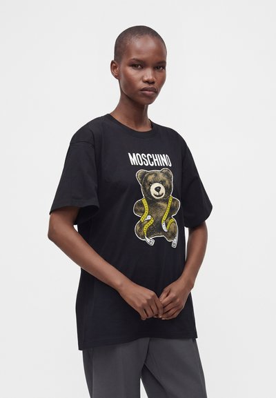 Άτομο με κοντά μαλλιά φοράει μαύρο T-shirt Moschino με γραφικό αρκουδάκι και κίτρινη μεζούρα σε απλό φόντο.