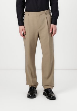 Pantalones beige ajustados con pliegues, hechos de una tela suave. Presenta trabillas para el cinturón y perneras estrechas, combinados con zapatos de cuero negros.
