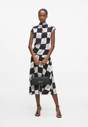 Donna che indossa un vestito midi a quadri bianco e nero con stampe schizzo, che tiene una borsa nera con scritta dorata “Karl” e tacchi neri con cinturino alla caviglia.