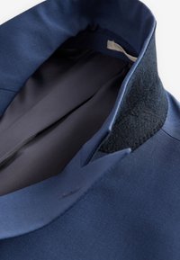 Col de veste bleu avec un patch texturé foncé et une doublure intérieure lisse, montré en gros plan sur fond blanc.