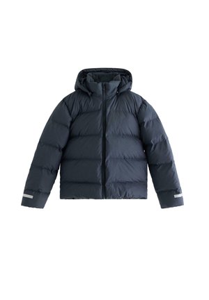 Giovane persona che indossa un berretto di lana marrone, una giacca imbottita blu scuro, una camicia a quadri bianca e nera, jeans scuri e sneakers bianche, in piedi con le mani in tasca.