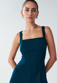 Obleka s kvadratnim izrezom v barvi modrozelene, z debelimi naramnicami in oprijetim zgornjim delom, izdelana iz strukturirane tkanine. Model nosi velike zlate obroče za ušesa.