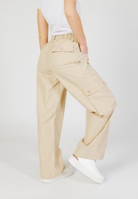 Pantaloni cargo beige con fascia elastica in vita, tasche laterali e tasche a patta con bottone. Tessuto liscio e design ampio a gamba larga.