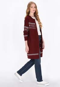 Hosszú bordó kardigán fehér és kék téli mintákkal, bordázott mandzsettákkal és elülső gombokkal, kék farmerrel és fehér tornacipővel párosítva.