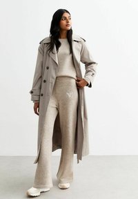 Manteau trench beige sur un haut en tricot beige et un pantalon évasé assorti, avec un logo brodé, le tout sur un fond clair.