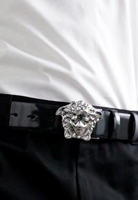 Curea din piele neagră cu o cataramă mare și complicată din silver în formă de față cu păr ondulat, așezată pe un fundal de cămașă albă.