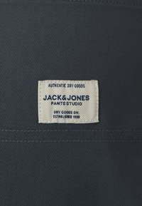 Étiquette pour un pantalon en tissu bleu avec un patch tissé sur fond blanc, texte bleu indiquant la marque, l'authenticité et l'année de création.
