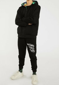 Personne portant un sweat à capuche noir avec une capuche intérieure colorée et un pantalon de survêtement noir assorti avec un texte graffiti blanc "Rien n'est facile".