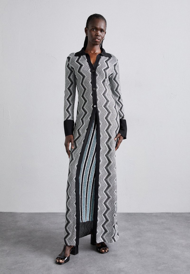 Ilgas juodos ir baltos zigzago rašto suknelė su juodu apykakle ir mygtukų uždarymu, turinti skeltą dizainą, kuris atskleidžia mėlynas ir baltas vertikalias juosteles.