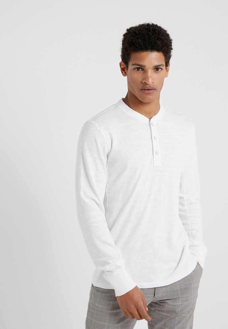 Hvit langermet henley-skjorte med rund hals og fire knapper foran. Har et mykt, teksturert stoff. Kombinert med rutete bukser.