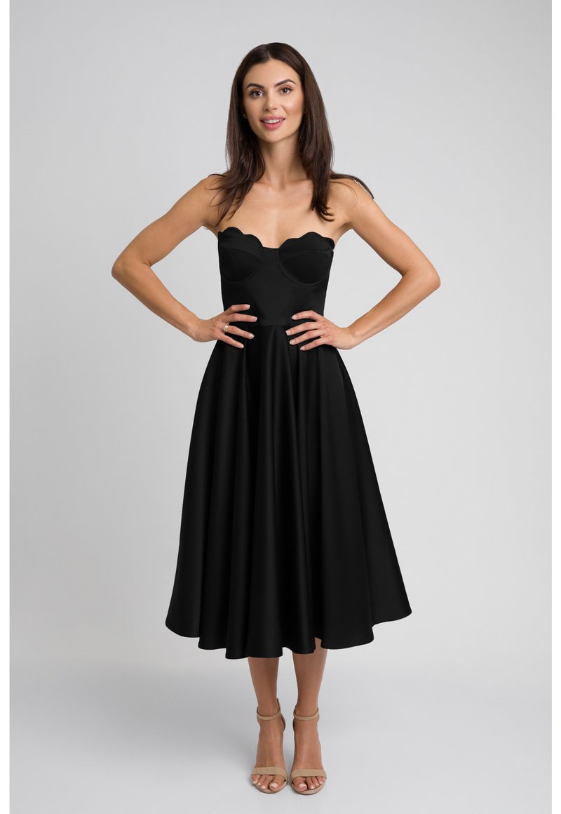 Czarna sukienka bez ramiączek z falistym dekoltem, dopasowaną górą i rozkloszowanym dołem. Gładki materiał i cieliste szpilki z paskiem na kostce.