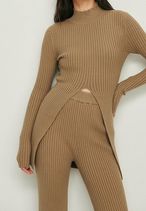 Kvinde iført matchende ribstrikket taupe sweater med sideslids og højtaljede bukser, stående mod en enkel baggrund.