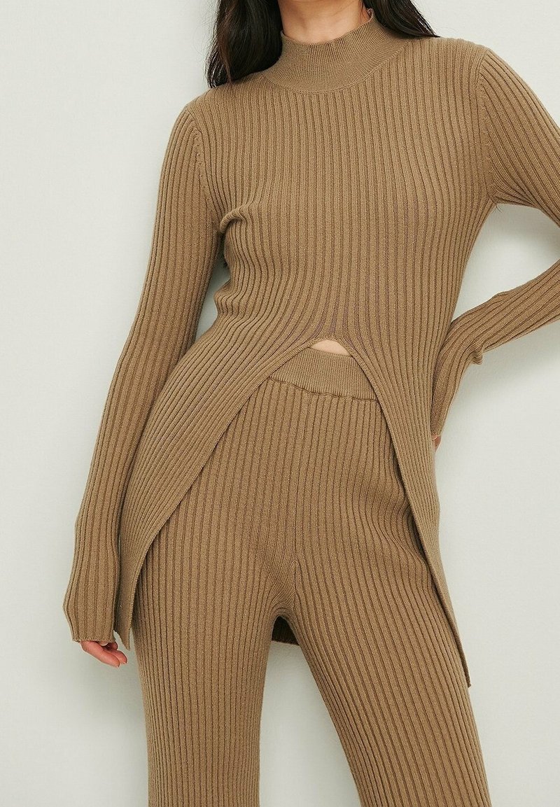 Femme portant un pull côtelé taupe assorti avec des fentes latérales et un pantalon taille haute, debout devant un fond uni.