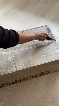 Kraftový kartónový box s priesvitnou páskou, čiastočne otvorený, s výrazným logom Zalando. Ruka s univerzálnym nožíkom reže pásku.
