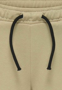 Beige sweatpants van een textuurstof, met een zwarte trekkoord en elastische tailleband. Stiksels zijn zichtbaar langs de naden.