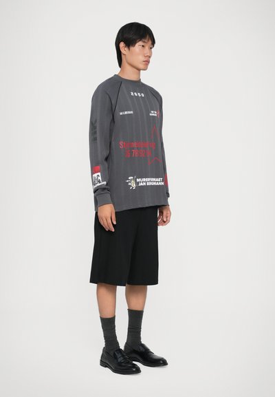 Jovem em pé, vestindo uma camisa de manga longa cinza com gráfico, calções pretos até ao joelho, meias cinzas e sapatos de couro pretos, em frente a um fundo simples.