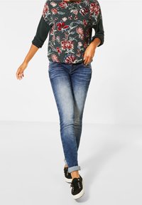 Floral-patterned dark green top with three-quarter sleeves, paired with fitted blue denim jeans and black sneakers with white soles.
