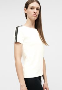 Γυναίκα με μακριά καστανά μαλλιά, φορώντας λευκό κοντομάνικο πουκάμισο με μαύρες ρίγες και κείμενο στους ώμους, στέκεται μπροστά από απλό φόντο.