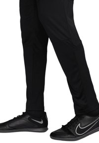 Pantalons de sport noirs avec une texture lisse et des jambes fuselées, associés à des baskets noires avec un swoosh Nike en argent et des accents texturés.
