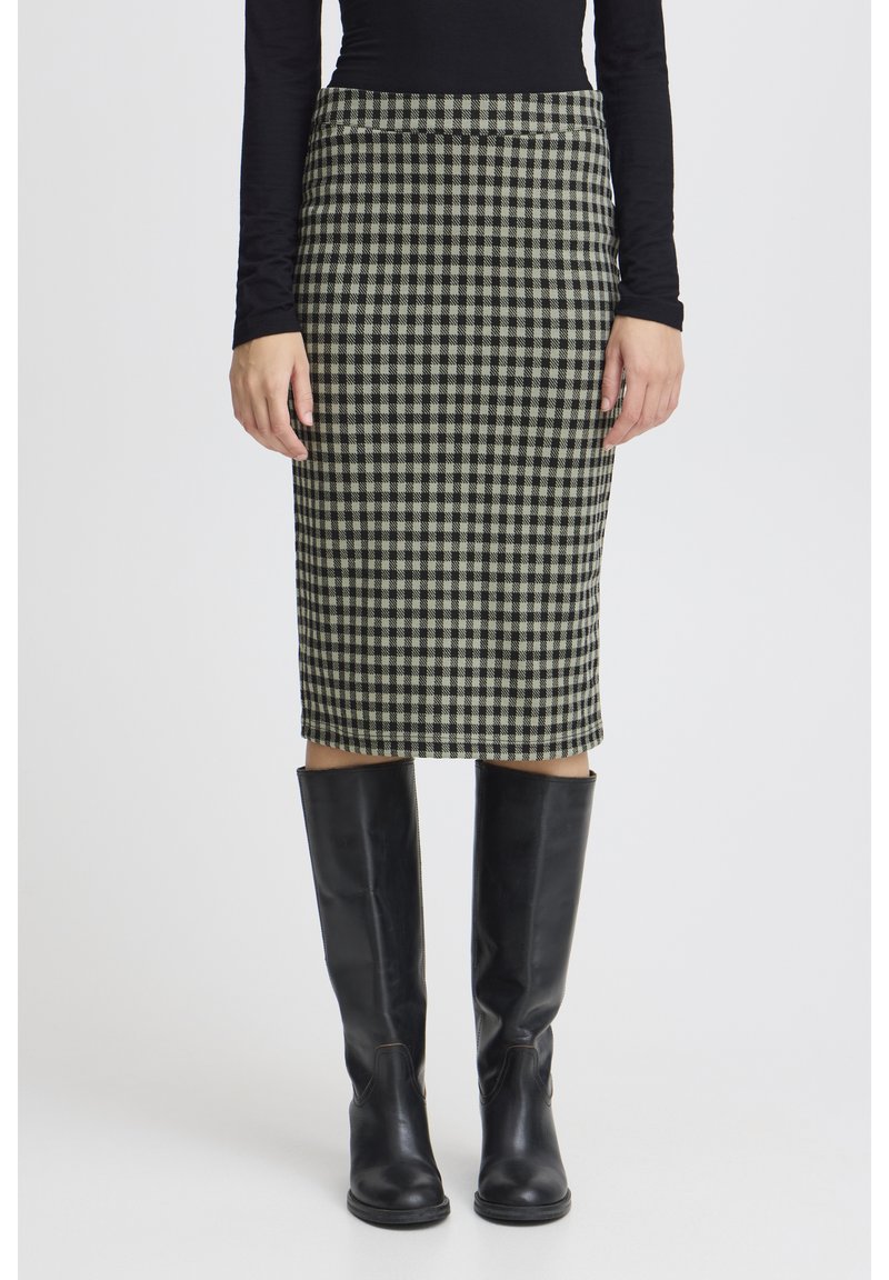 Woman wearing a black long-sleeve top, black and white checkered pencil skirt, and black knee-high boots standing against a plain background.