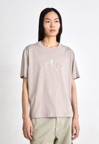 Šviesiai kreminės spalvos medvilnės marškinėliai su apvaliomis iškirptėmis ir trumpomis rankovėmis. Papuošti siuvinėtų "CK" logotipu ir subtiliu tekstu "Calvin Klein Jeans".