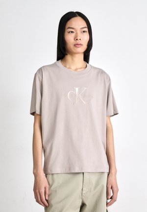 Jasno beżowa bawełniana koszulka z okrągłym dekoltem i krótkimi rękawami. Posiada haftowane logo „CK” oraz napis „Calvin Klein Jeans” w subtelnej formie.