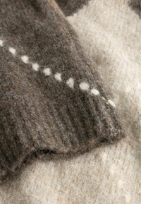 Close-up van zachte gebreide stof met een geribde bruine mouw met witte hartvormige patronen op een beige, gestructureerde achtergrond.