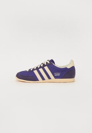 Purple suede sneaker with perforations, three beige stripes, and cream-colored accents. Features a rubber sole and branding on the side.