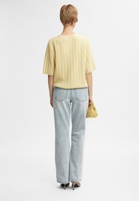 Person med kort hår som bär en gul kortärmad ribbad tröja, ljusblå jeans, beige klackar, håller en liten gul väska och vänder ryggen till.