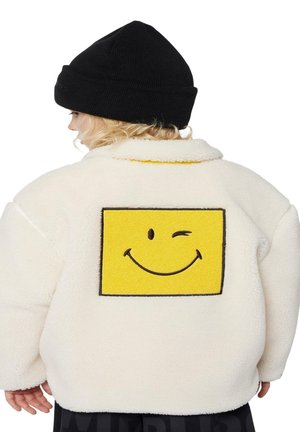 Veste en polaire blanche avec un grand patch de smiley jaune au dos. Béret en tricot noir visible sur la tête, avec un tissu texturé.