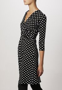 Svart klänning med vita prickar, V-ringning, trekvartslånga ärmar, figursydd midja och A-linjefjäril. Tillverkad av mjukt, elastiskt tyg.