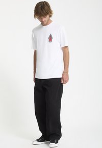 Hvid bomulds T-shirt med korte ærmer, prydet med et farverigt grafisk design på brystet. Bæres med løstsiddende sorte bukser og sorte sneakers.