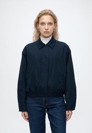Femme aux cheveux blonds portant une veste boutonnée bleu foncé sur un haut blanc et un jean bleu, debout devant un fond clair uni.