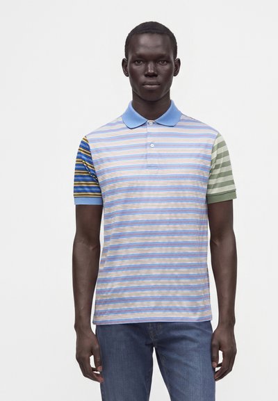 Homme portant un polo bleu à col avec des rayures horizontales à motifs variés et un jean bleu, debout devant un fond blanc uni.