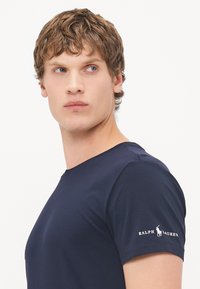 Tricou din bumbac de culoare albastră închis, cu guler rotund și mâneci scurte, având un logo alb "Ralph Lauren" și un emblema cu jucător de polo pe mâneca stângă.