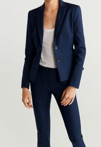 Tmavě modrý oblek na míru s jednoklapkovým sakem a úzkými kalhotami, oboje vyrobené z hladké látky, s jemnými knoflíkovými detaily.
