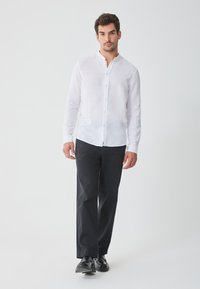 Witte Linnen overhemd met lange mouwen, met knoopsluiting kraag, gedragen met donkere broeken en zwarte schoenen. Eenvoudig, schoon ontwerp en lichte stof.