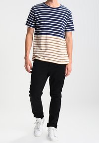 Pruhované tričko s krátkým rukávem v námořnické modré a béžové barvě, s horizontálními pruhy, kulatým výstřihem a jemnou texturou. Doplněno černými kalhotami a bílými teniskami.